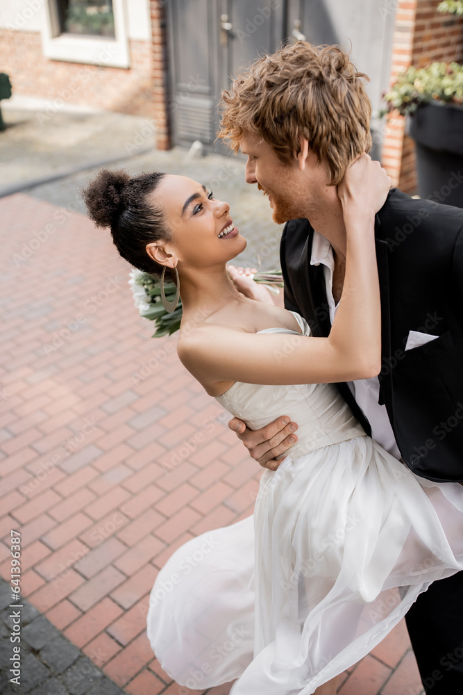 overjoyed and elegant interracial couple in wedding attire embracing on urban street
