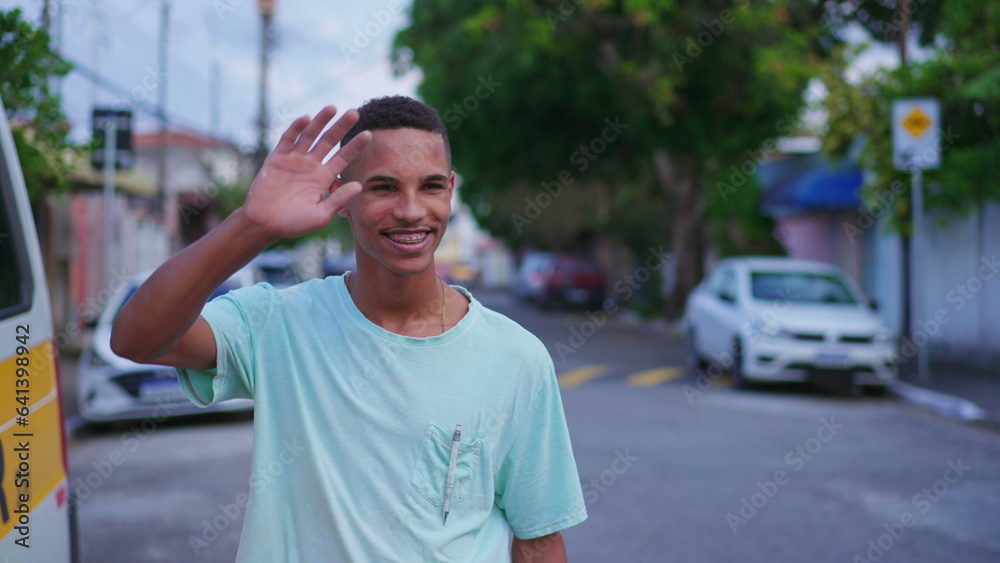 One cheerful young Brazilian man waving hello to neighbor walking in street. Joyful South American individual greeting friend with hand
