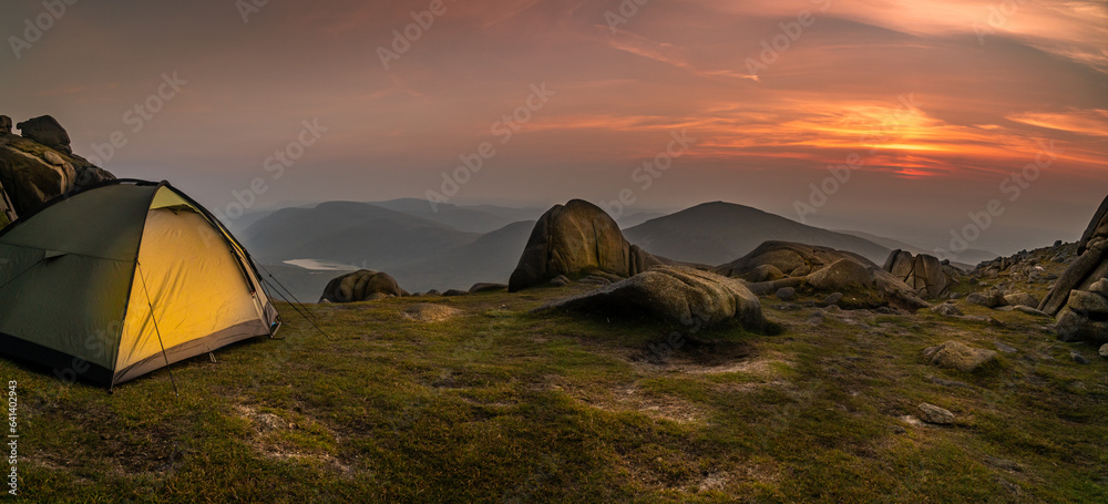 Glowing orange and green tent in the mountains under dramatic evening sky. Red, purple sunset and mountains in the background. Summer landscape. Panorama. Lake in the background.