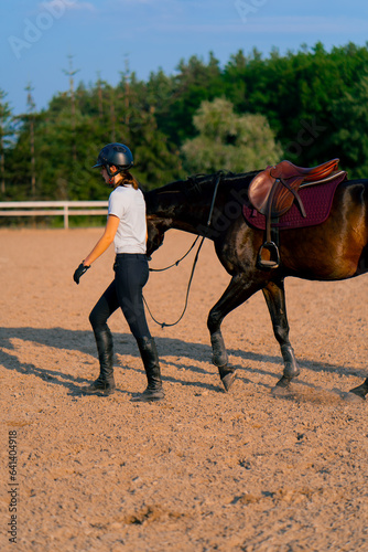 A helmeted rider leads her beautiful black horse by the harness in the riding arena during a horseback ride