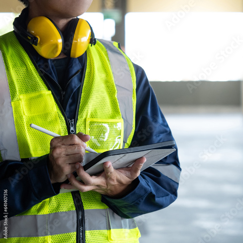 Building inspector man using digital tablet checking safety and security system. Asian male worker in reflective vest and protective ear muffs working for building maintenance inspection