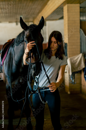 A rider puts harness on her black horse in the stables in preparation for a race equestrian concept 