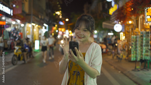 Portrait of happy Asian Vietnamese woman travel at night market, using mobile phone in technology device in people walking street fair in Hanoi city, Vietnam. Retail shops