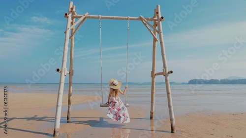 Girl relaxing on beach swing with tropical sea view. Woman enjoying summer vacation in a serene and beautiful setting. Concept of peaceful beach getaway. Back view, slow motion.
