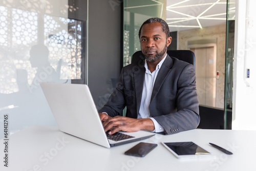Portrait of serious thinking and confident businessman, african american boss thinking and looking at camera, man working inside office in business suit, investor financier in with laptop.