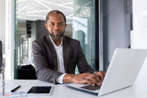Portrait of serious thinking and confident businessman, african american boss thinking and looking at camera, man working inside office in business suit, investor financier in with laptop.