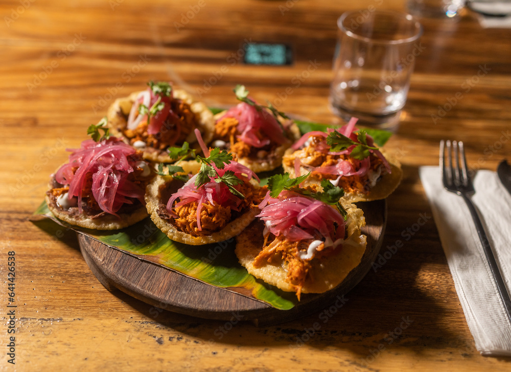 Traditional Mexican cuisine with tortilla and red onions served on top of banana leaf in a wooden plate with transparent glass in the background