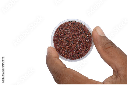 Overhead view of the tiny red Garnet mineral pieces filled on a small round tray is held by the two fingers in isolated background