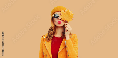 Autumn color style outfit, portrait of stylish beautiful young woman model with yellow maple leaves blowing her lips sends sweet kiss wearing orange french beret, sunglasses on brown studio background