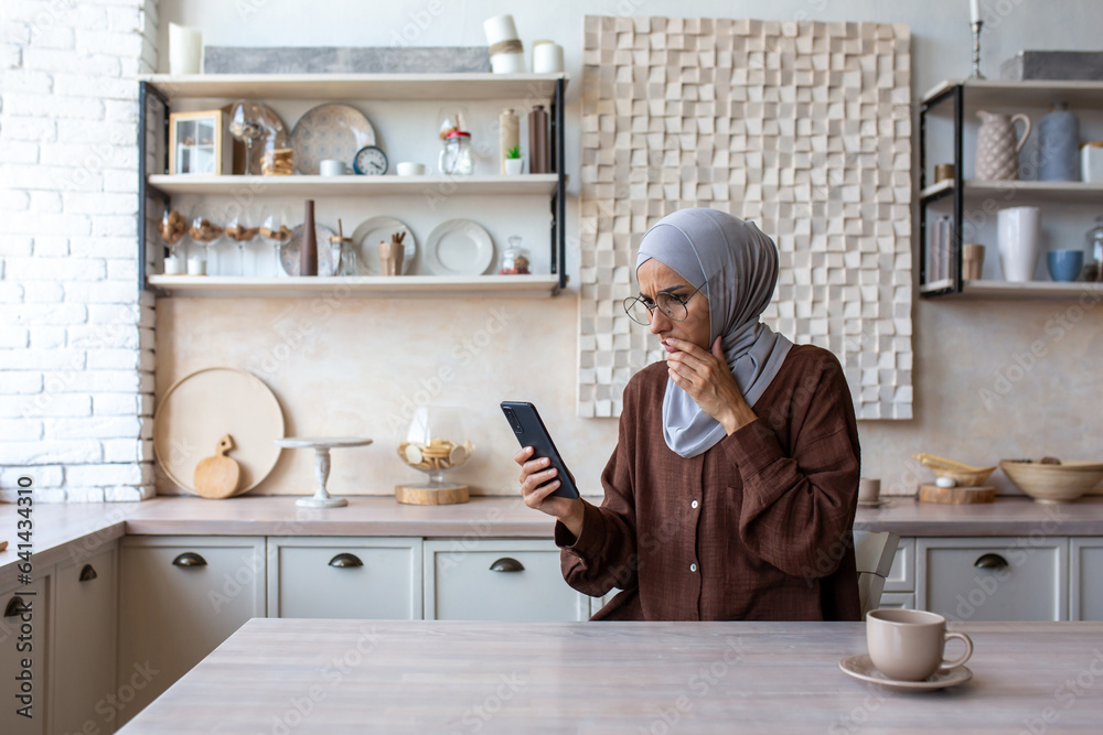 Φωτογραφία Muslim young woman wearing hijab sitting at home in kitchen and looking shocked at phone, covering mouth with hand reading message and bad news