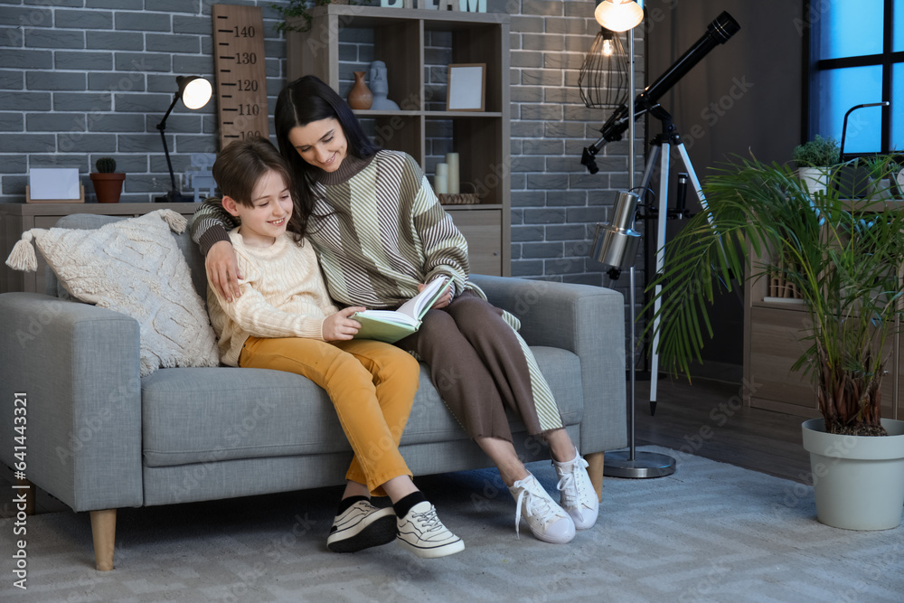 Fototapeta premium Little boy with his mother sitting on sofa and reading book at home late in evening