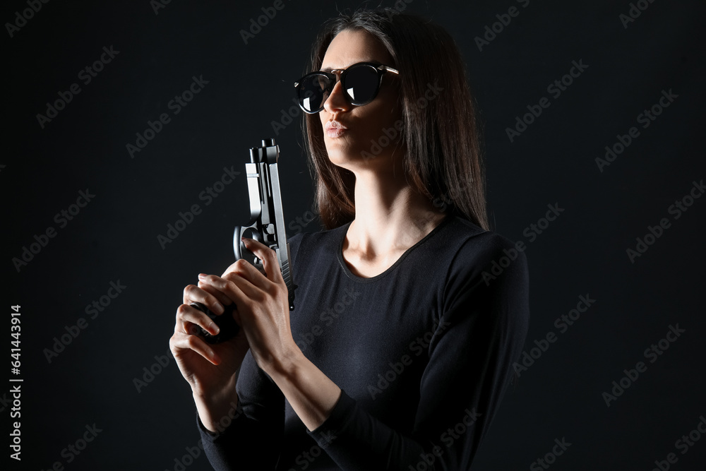 Stylish young woman in sunglasses with gun on black background