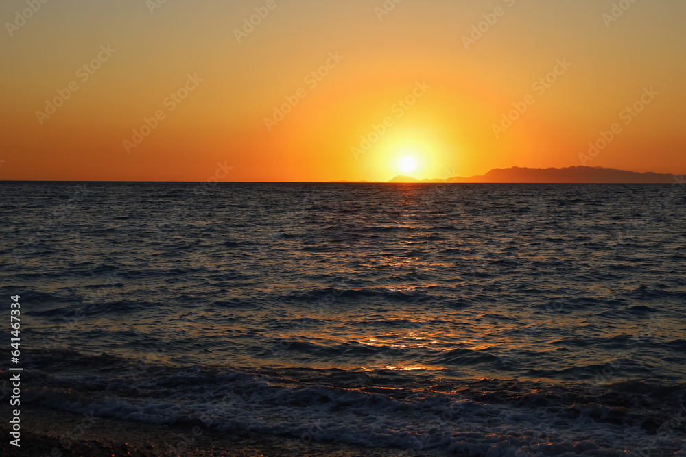 Colorful vibrant orange sunset on the seaside. Golden hour sunlight. Majestic dusk. Seascape in Rhodes, Greece. Reflection of sun path on horizon. Abstract nature and travel background