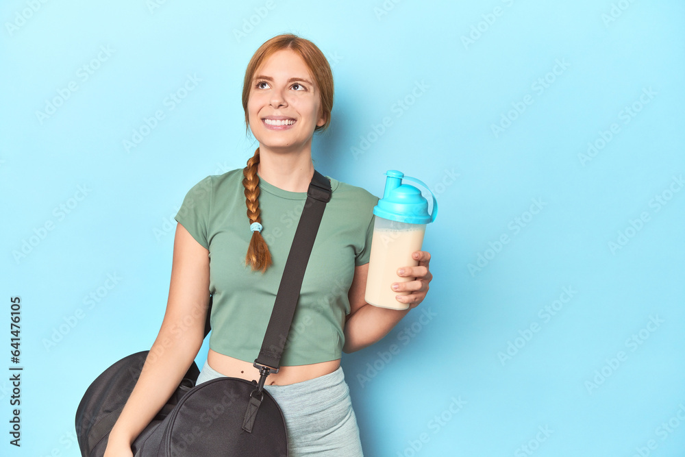 Redhead holding protein shake in blue studio dreaming of achieving goals and purposes Redhead holding protein shake in blue studio dreaming of achieving goals and purposes