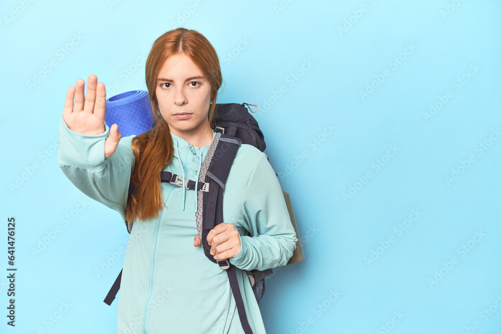 Redhead traveler with backpack and mat in studio standing with outstretched hand showing stop sign, preventing you. Redhead traveler with backpack and mat in studio standing with outstretched hand showing stop sign, preventing you.