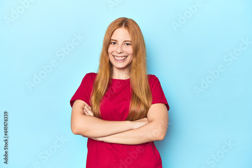 Redhead young woman on blue background who feels confident, crossing arms with determination. © Asier