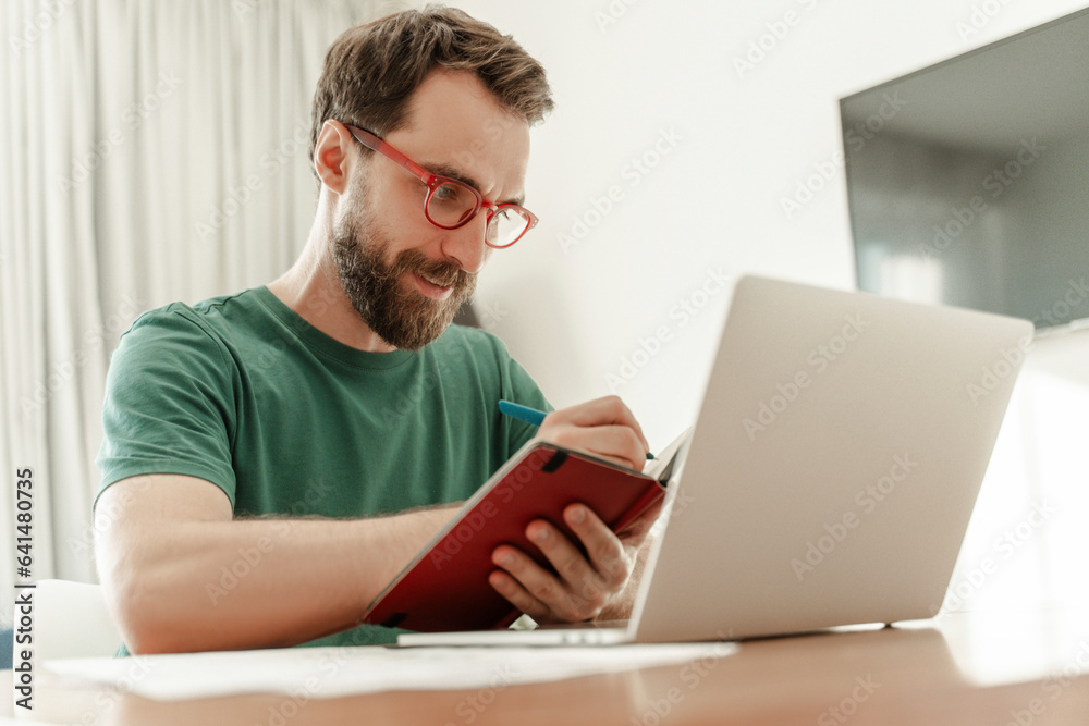 Bearded writer holding notebook and pen, using laptop, sitting at workplace at home, taking notes Bearded writer holding notebook and pen, using laptop, sitting at workplace at home, taking notes
