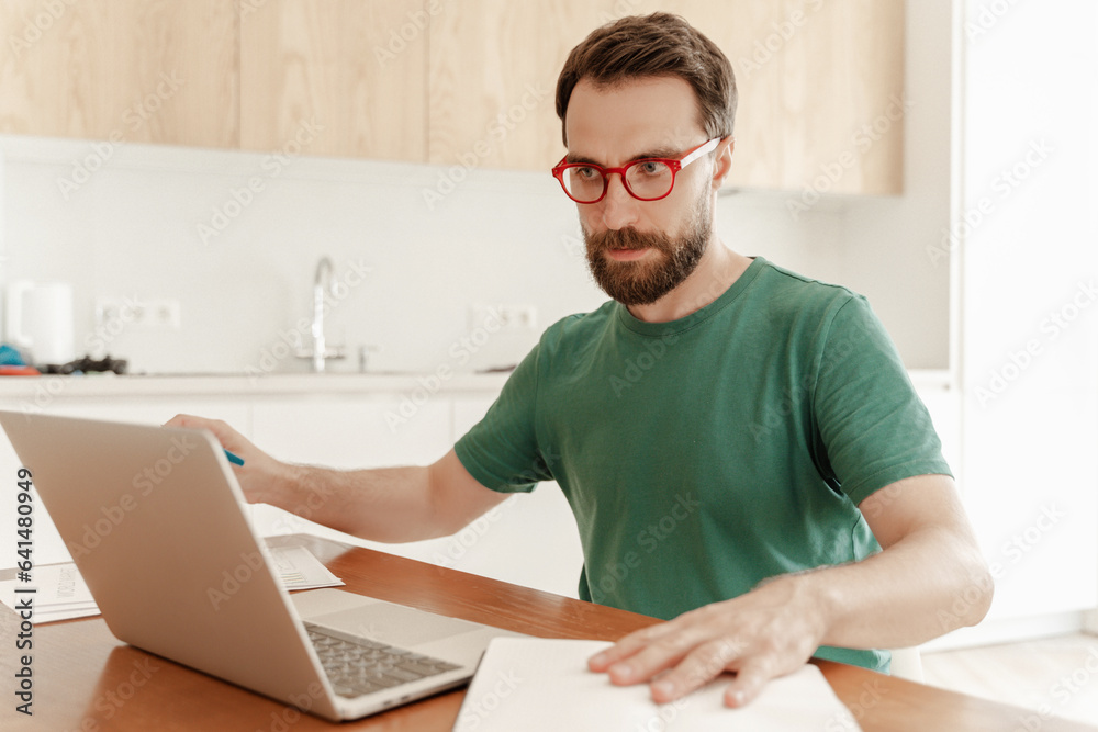 Pensive male student wearing red glasses and casual green t shirt using laptop, preparing for exam Pensive male student wearing red glasses and casual green t shirt using laptop, preparing for exam