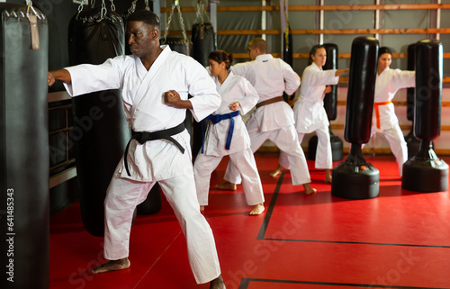 African-american man and group of people in kimono exercising with punching bags in gym during karate training.