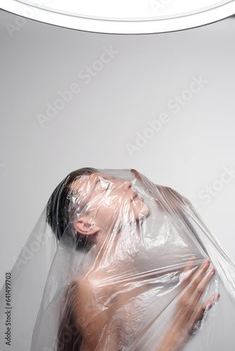 Side view of naked young man suffocating wrapped in plastic sheet against white wall background  photo