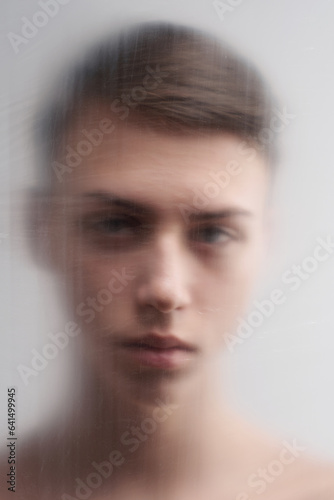 Portrait of gloomy brown-haired male teenager standing behind plastic film and looking at camera 