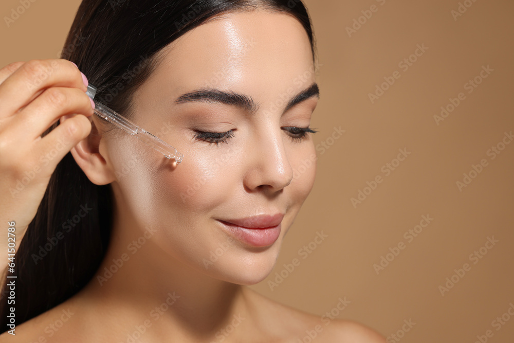 Beautiful young woman applying serum onto her face on beige background, closeup. Space for text