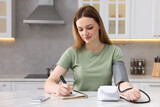 Woman measuring blood pressure and writing it down into notebook in kitchen, space for text