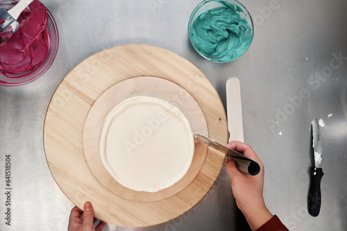 Top view of unrecognizable pastry chef coating cake with white cream, bowls with colorful toppings seen on table beside  photo