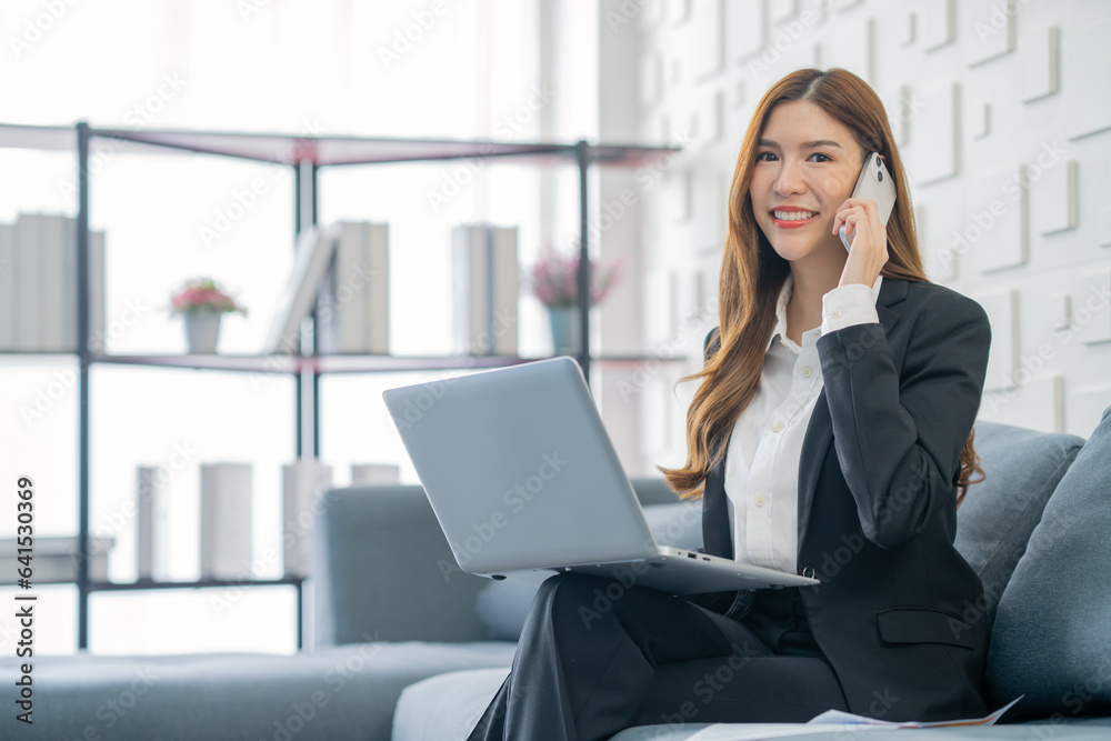 Image of a young beautiful Asian Business woman smiling while working with a laptop and talking to work doing math finance on an office desk, tax, report, accounting, statistics, and research concepts Image of a young beautiful Asian Business woman smiling while working with a laptop and talking to work doing math finance on an office desk, tax, report, accounting, statistics, and research concepts