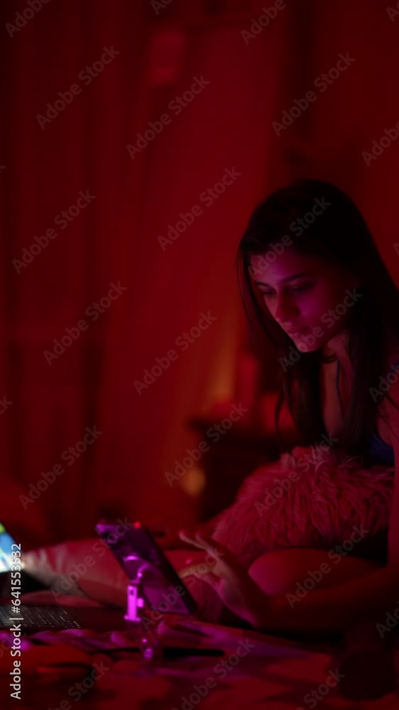 A young woman is working on her laptop while in a dimly lit bedroom, seated on the bed. A young woman is working on her laptop while in a dimly lit bedroom, seated on the bed.