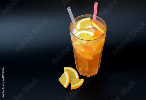 A tall faceted glass of refreshing lemonade with ice on a black background, next to pieces of a ripe orange.