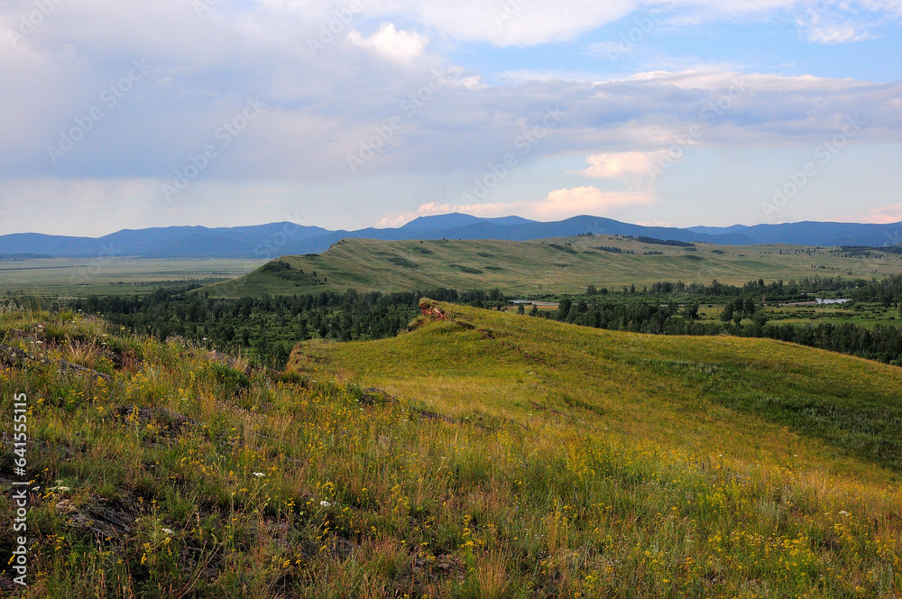 View from the top of the hill at the mountain range in the endless steppe and the meandering river flowing in the lowland under a cloudy summer sky.