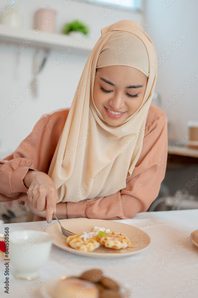 A beautiful Asian Muslim woman eating waffles, having her breakfast at a table in the kitchen A beautiful Asian Muslim woman eating waffles, having her breakfast at a table in the kitchen