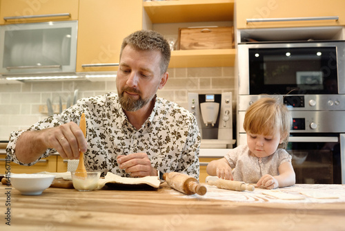 Adult bearded man and little charming girl at table in kitchen making sweet pastries to bake