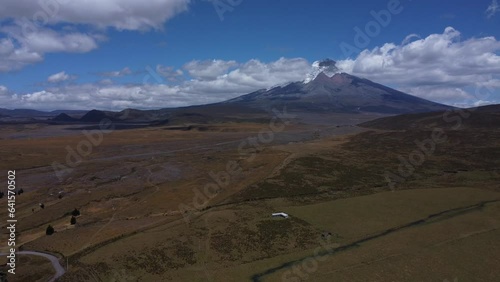 Capture the grandeur of Cotopaxi Volcano in 4K drone footage, as our camera smoothly pans across the majestic peak on a sunny day, framed by the blue sky and adorned with gentle cloud veils