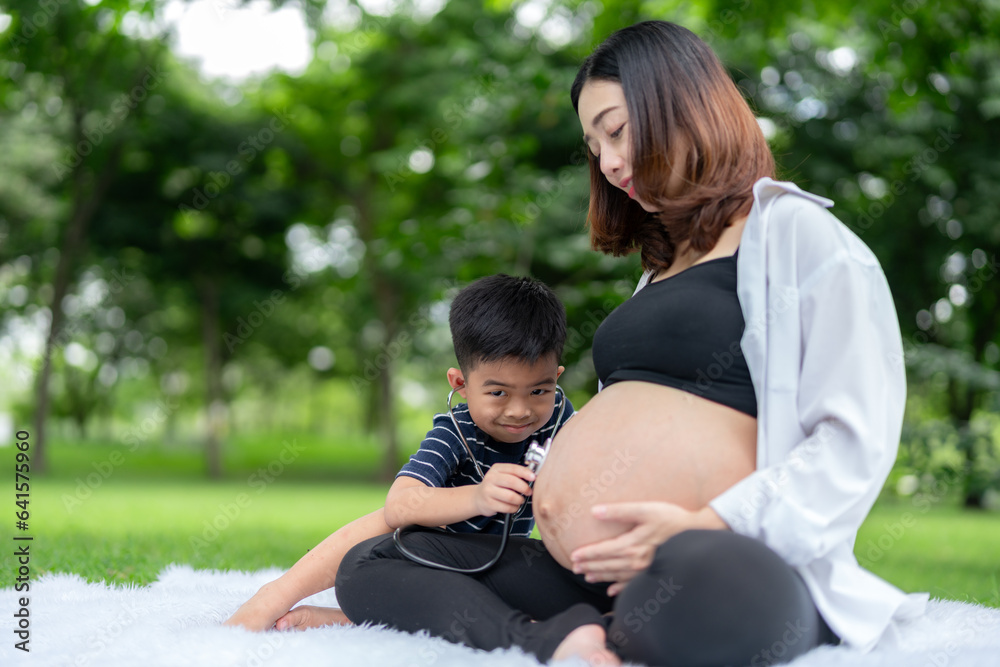 Portrait of a pregnant woman relaxing in the park with her son. Older brother uses headphones to listen to his younger brother in the womb Portrait of a pregnant woman relaxing in the park with her son. Older brother uses headphones to listen to his younger brother in the womb