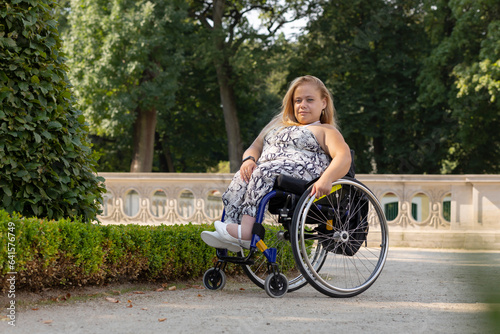 Full Length Young Pretty Woman With Short Stature On Wheelchair Looking At Camera in Public Park At Summer Day. Smiling Pretty Female Adult With Disability. Horizontal Plane photo