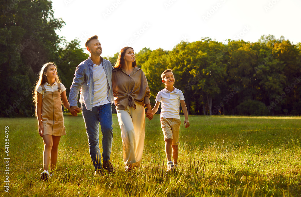 Fototapeta premium Parents and children take a stroll in nature. Happy mother, father and children holding hands and walking together on a grass lawn in a beautiful green park on a good sunny summer day. Family concept