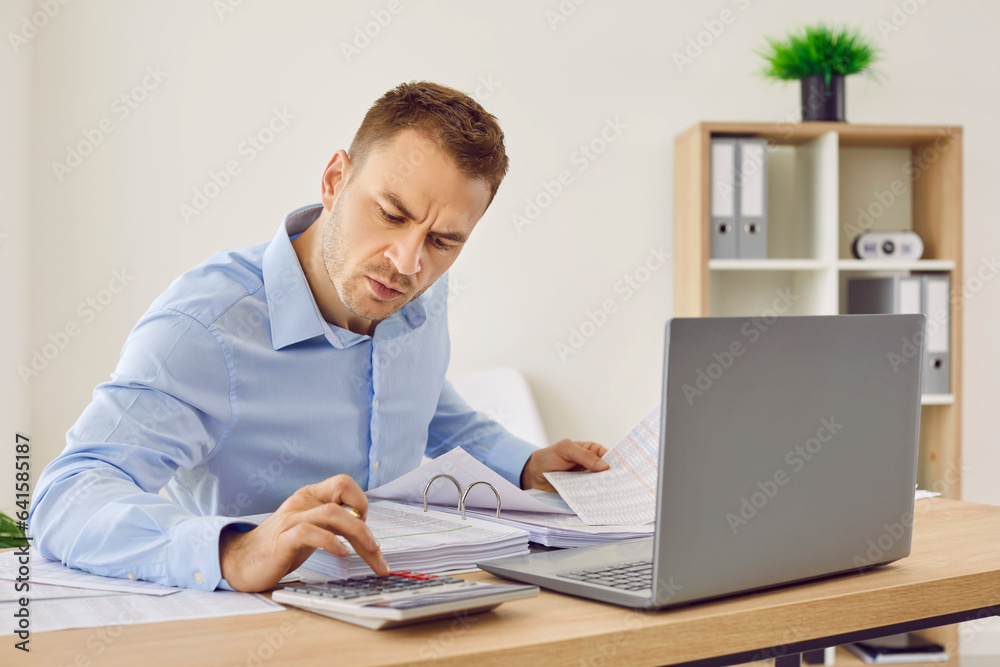 Financial accountant calculating something in office. Man in blue shirt working in office, sitting at his desk, using laptop and calculator, and working with papers. Finance, business, budget concept Financial accountant calculating something in office. Man in blue shirt working in office, sitting at his desk, using laptop and calculator, and working with papers. Finance, business, budget concept