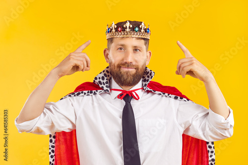Portrait of a handsome smiling bearded young cheerful man wearing white shirt with a tie with crown of a king and mantle looking confidently at the camera isolated on a studio yellow background. photo