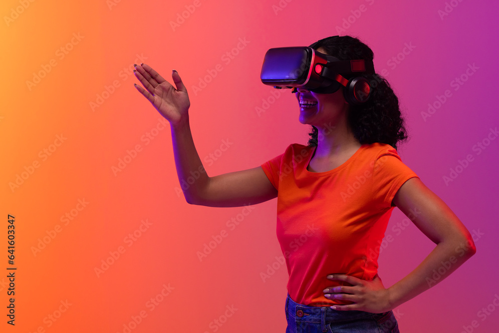 Biracial woman using vr headset and pointing on neon pink to orange background Biracial woman using vr headset and pointing on neon pink to orange background