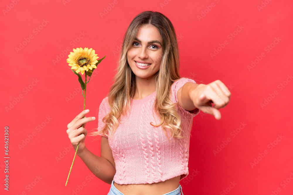 Young pretty Uruguayan woman holding sunflower isolated on background points finger at you with a confident expression Young pretty Uruguayan woman holding sunflower isolated on background points finger at you with a confident expression