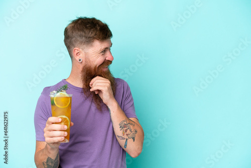 Redhead man with long beard holding a cocktail isolated on blue background thinking an idea and looking side