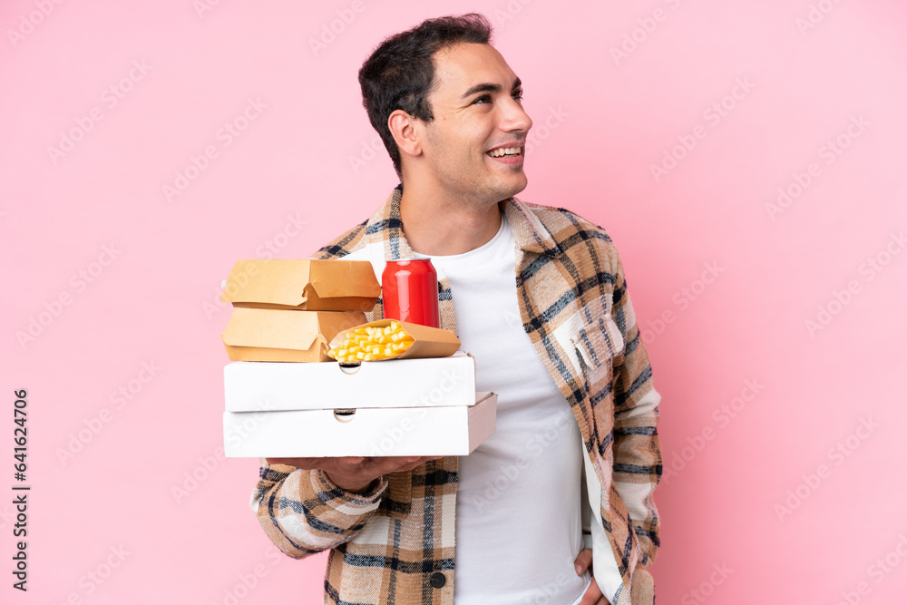 Young caucasian man holding fast food isolated on pink background posing with arms at hip and smiling Young caucasian man holding fast food isolated on pink background posing with arms at hip and smiling