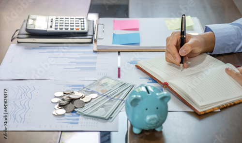 Businessman hand writing income and expenses to account with piggy bank and dollar notes, step up start-up businesses to success, Saving money for future plan and retirement fund concept