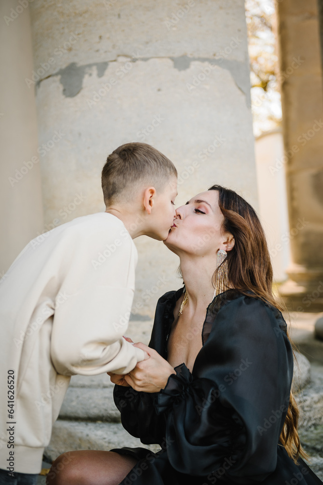 Mother hold hands son stands on stairs near large columns of ancient temple at sunset. Parent kiss kid boy walk on autumn day. Child hugs mom near Baroque Catholic church in Pidhirtsi Lviv Ukraine