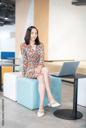 Portrait of an Asian female executive with short hair company head Wear a floral suit Man looking at camera near business laptop on sofa chair In the office space There is a large monitor at the back.