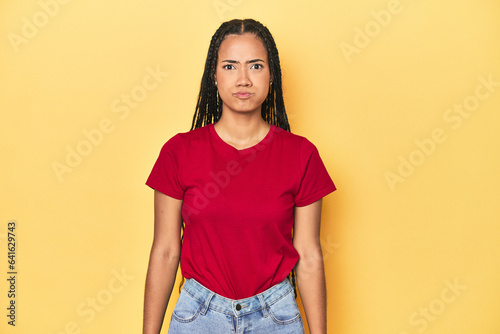Young Indonesian woman on yellow studio backdrop blows cheeks, has tired expression. Facial expression concept.