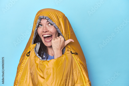 Woman in sleeping bag on blue background points with thumb finger away  laughing and carefree.