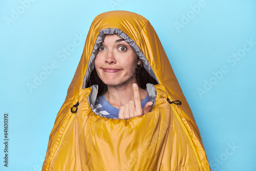 Woman in sleeping bag on blue background pointing with finger at you as if inviting come closer.